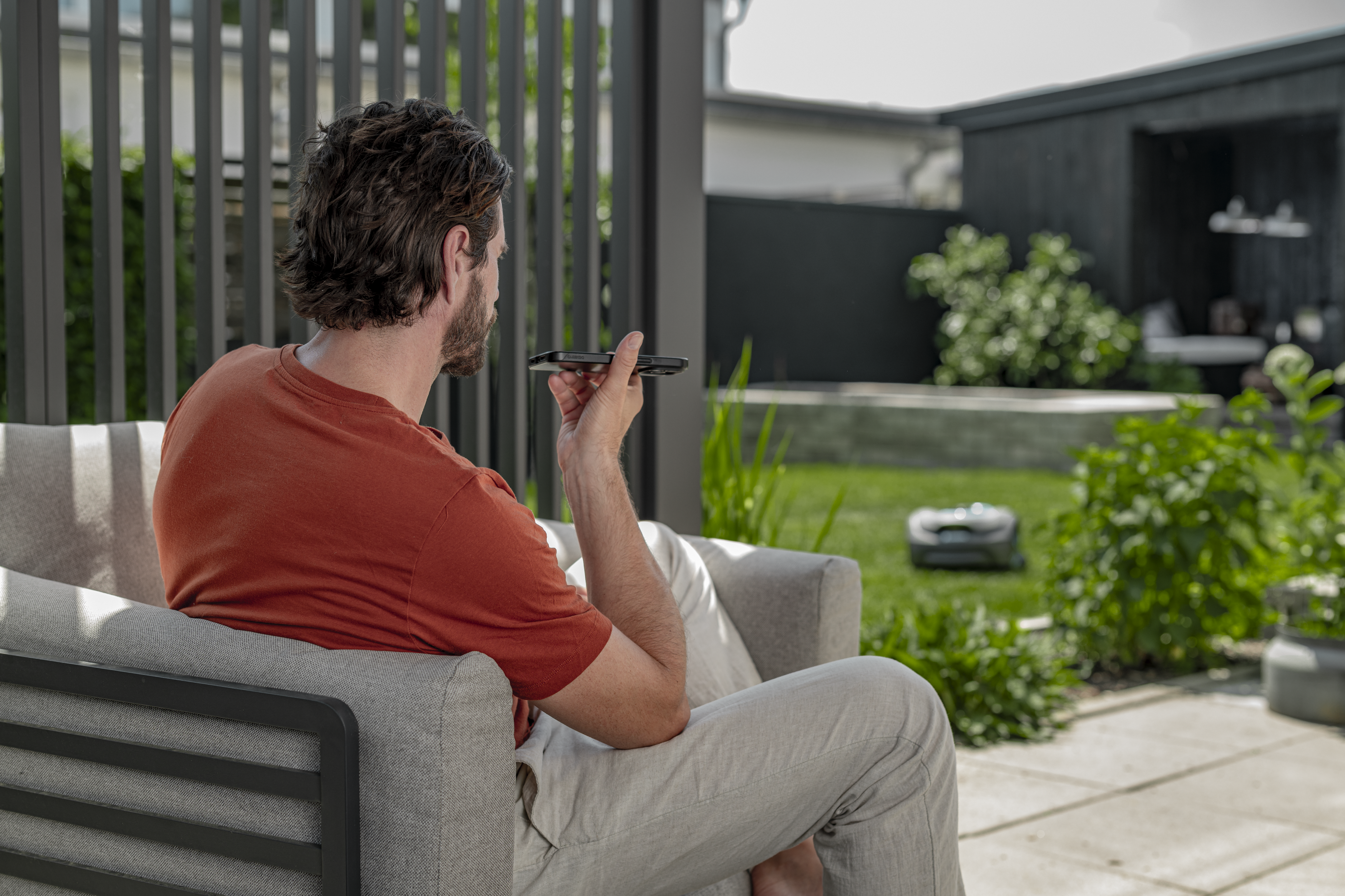 Person seated on a patio sofa using a smartphone, with a garden and house in the background.
