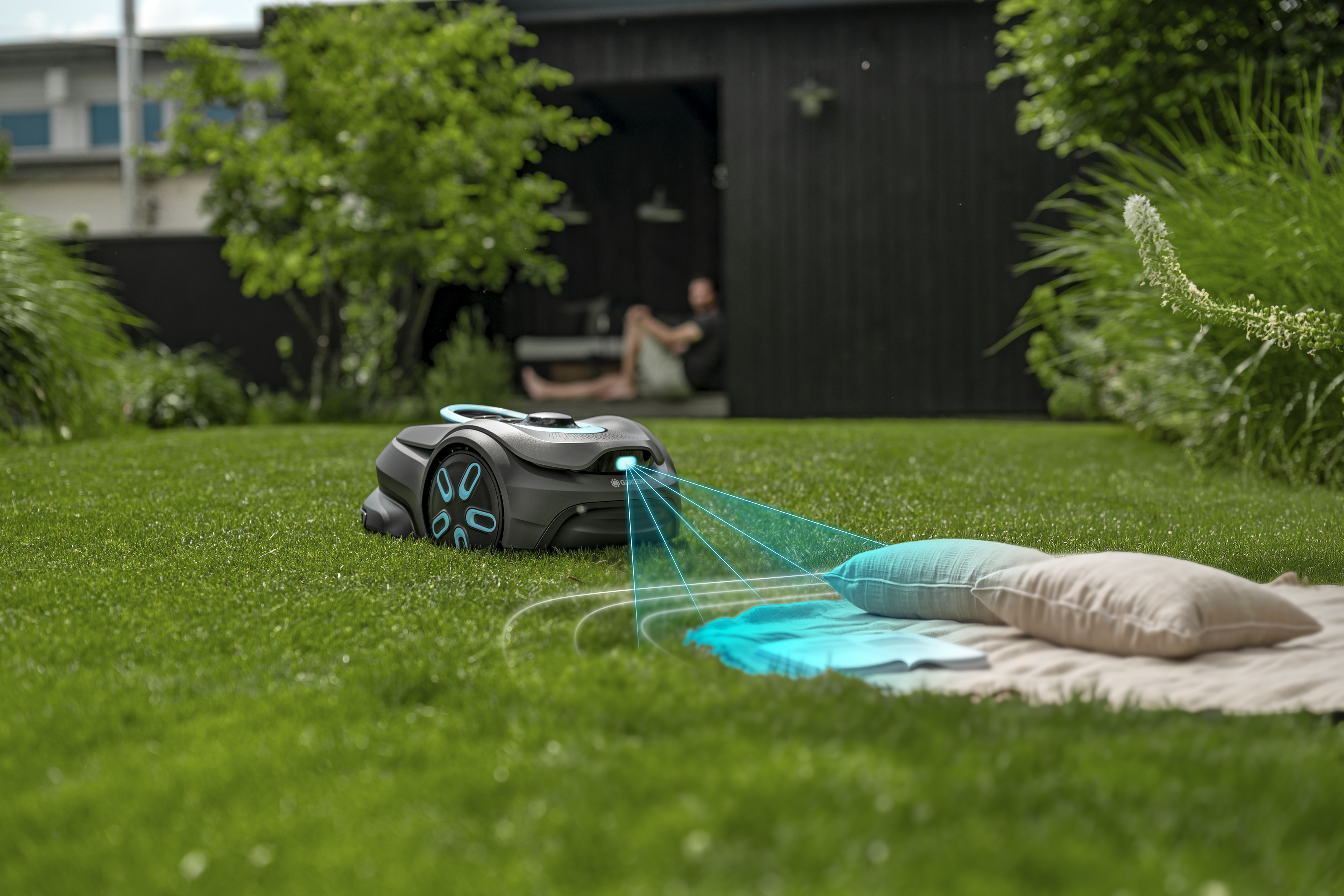 Robotic lawn mower on a garden lawn near a blanket with pillows, with house and greenery in the background.